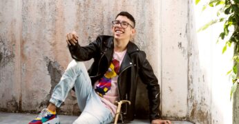 Imagem de Ivan Baron. Ele é um homem com cabelos curtos e óculos. Ele usa calça jeans com tonalidade clara, jaqueta preta e uma camiseta branca com uma estampa frontal. Ivan está sentado em um banco, no qual uma bengala está apoiada.