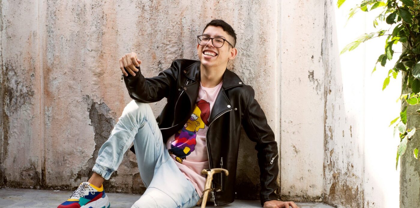 Imagem de Ivan Baron. Ele é um homem com cabelos curtos e óculos. Ele usa calça jeans com tonalidade clara, jaqueta preta e uma camiseta branca com uma estampa frontal. Ivan está sentado em um banco, no qual uma bengala está apoiada.