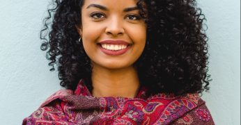 Trata-se da foto de Lígia Batista. Ela tem cabelos encaracolados e usa uma echarpe com tons de magenta. Ela aparece sorrindo na imagem.