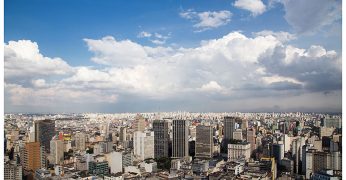 Imagem do horizonte da região central de São Paulo. A imagem é composta por um conjunto de construções, com destaque para prédios, e é possível ver o céu ensolarado.