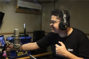 Imagem de Caio Santos. Ele tem cabelos curtos, usa óculos e veste uma camiseta na cor preta. Ele usa também fones de ouvidos durante a gravação de um podcast.