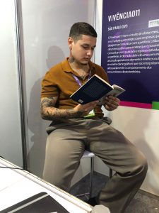 Imagem de Wesley Xavier. Wesley aparece lendo um livro. Ele tem cabelos curtos, uma tatuagem no lado direito do pescoço e veste calças beges e uma camisa manga curta amarela em tonalidade mostarda.