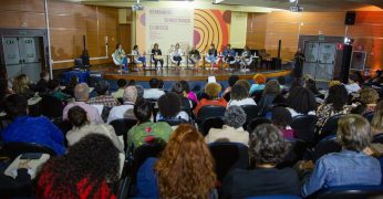 Na imagem, o auditório da EACH-USP aparece lotado durante a abertura do Seminário Territórios Clínicos. Aparecem oito pessoas no palco, em frente ao toldo com a divulgação do evento.