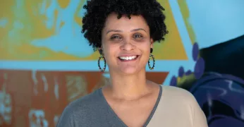 Imagem de Viviane Soranso. Viviane é uma mulher negra, tem cabelos cacheados em formato black power, usa brincos em formato de argola e usa uma blusa bicolor - cinza e ocre. A imagem de Viviane, que aparece sorrindo e à frente de uma parede azul com desenhos, foi escolhida para ilustrar o post no qual ela indica cinco livros para quem quer se engajar na luta antirracista.