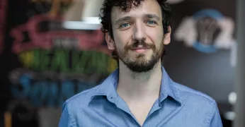 Foto de Pedro Marin. Ele tem barba, cabelos cacheados curtos e usa uma camisa azul. Ele aparece à frente de um fundo composto por uma parede preta com desenhos e de luminárias. A foto foi escolhida para ilustrar a publicação sobre cinco livros que falam sobre orçamento público.