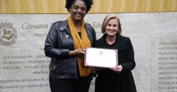 Imagem de Elaine Mineiro, vereadora do Quilombo Periférico, e Marleide Rezende, das Guardiãs do Território. Elas seguram a placa do Prêmio Chico Xavier de Reconhecimento Humanitário.