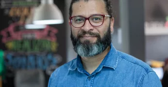 Imagem de Marcelo Ribeiro, coordenador de Prática de Desenvolvimento Local. Ele tem barba, cabelos longos presos e usa óculos e camisa na cor azul. Marcelo está à frente de um espaço composto por uma parede preta com desenhos e de luminárias. A foto foi escolhida para ilustrar o post sobre cinco obras para pensar garantia de direitos e desenvolvimento das periferias.