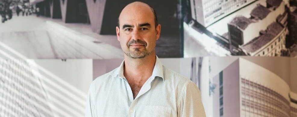 Imagem de Cesar Zucco. Ele usa uma camisa branca e tem o cabelo raspado. Ele está à frente de um fundo composto por um mural com fotos em preto e branco.