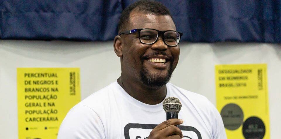 Imagem de Delton Aparecido Felipe. Ele é um homem negro. Ele veste uma camiseta branca, usa óculos na cor preta e segura um microfone.