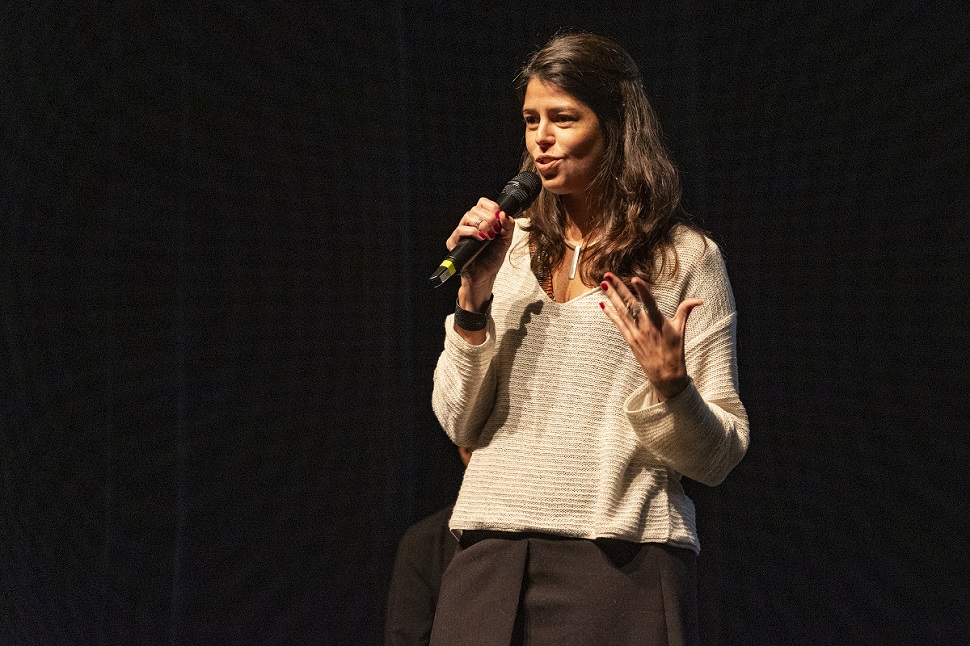 Imagem de Mariana Almeida durante o evento Plataforma Alas em Debate. Mariana fala ao microfone. Ela tem cabelos escuros longos, usa uma blusa branca e uma saia na cor preta.