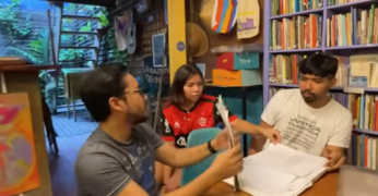 Cena do documentário 'Da Terra ao Clima'. A imagem mostra três pessoas (dois homens e uma mulher) sentados em uma mesa à frente de uma estante com livros e de uma área com jardim vertical ao fundo.