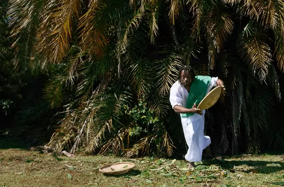 Cena da videodança 'De Bará a Oxalá'. A imagem mostra um artista caracterizado como Oxóssi e faz apresentação com balaio e flores em meio a uma área arborizada.