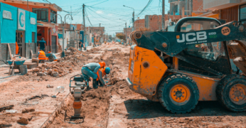 Imagem de obra infraestrutural para pavimentação de rua. Há operários trabalhando em uma via a ser asfaltada e na qual as calçadas serão readequadas. Há uma escavadeira à direita da foto.