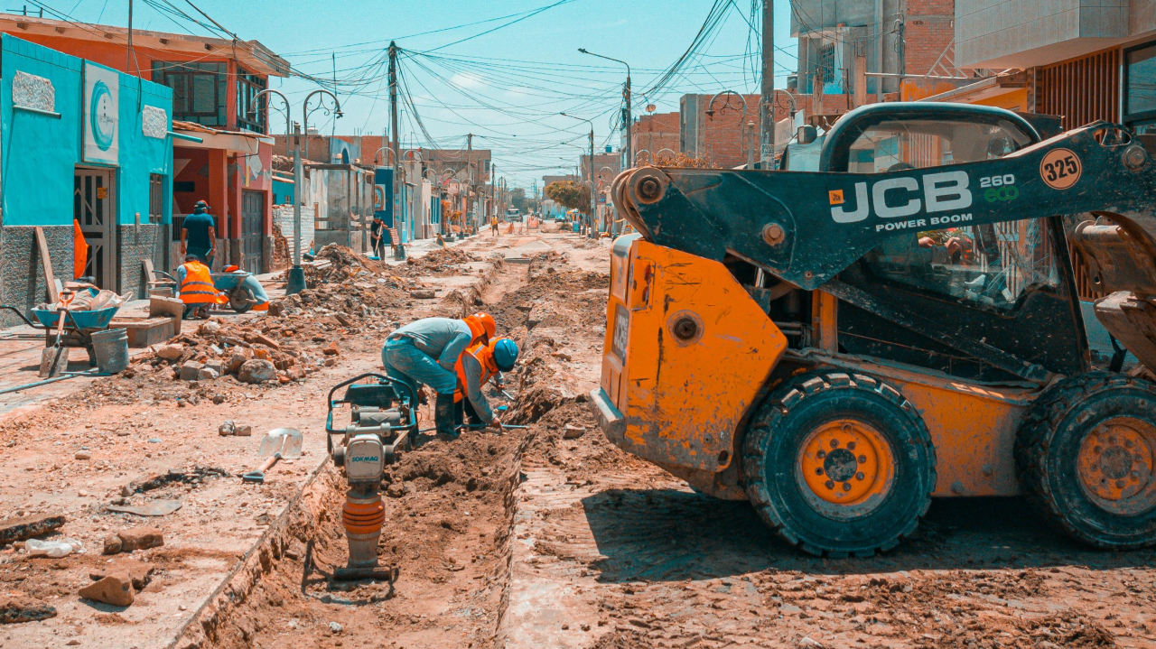 Imagem de obra infraestrutural para pavimentação de rua. Há operários trabalhando em uma via a ser asfaltada e na qual as calçadas serão readequadas. Há uma escavadeira à direita da foto.