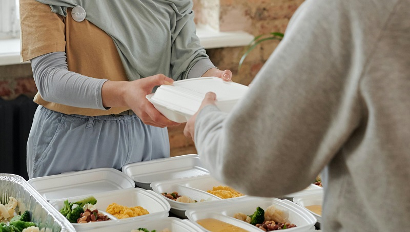 Imagem de uma pessoa entregando uma marmita para outra. Há outras marmitas sobre uma mesa. A foto ilustra o texto sobre insegurança alimentar.