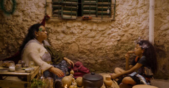 Cena do curta-metragem e documentário 'Onde a Terra Ferve'. A imagem mostra uma mulher à esquerda e uma criança à direita. Elas estão sentadas em cada canto, em frente à parede de uma casa, representando um contexto distópico.