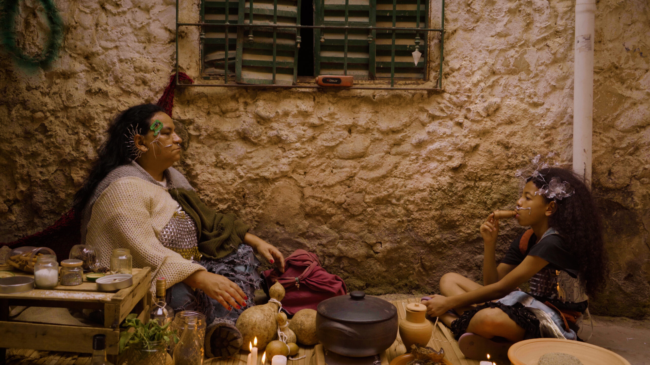 Cena do curta-metragem e documentário 'Onde a Terra Ferve'. A imagem mostra uma mulher à esquerda e uma criança à direita. Elas estão sentadas em cada canto, em frente à parede de uma casa, representando um contexto distópico.