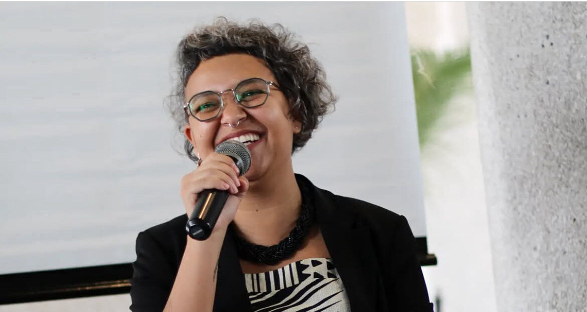 Foto de Fernanda K. Martins. Ela aparece falando ao microfone. Fernanda tem cabelo curto na altura do pescoço e encaracolado, usa blazer e colar pretos e vestido nas cores branco e preto.