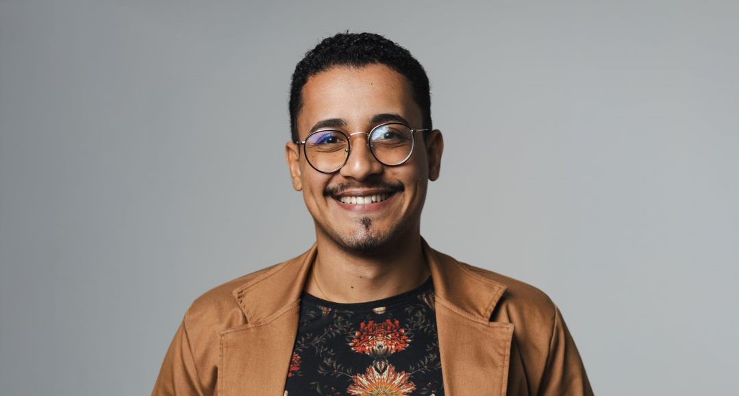 Imagem de Henrique Moreira. Ele é um homem negro com pele menos escura, cabelos curtos e barba. Ele usa óculos, um blazer na cor marrom e uma camiseta estampada na cor preta.