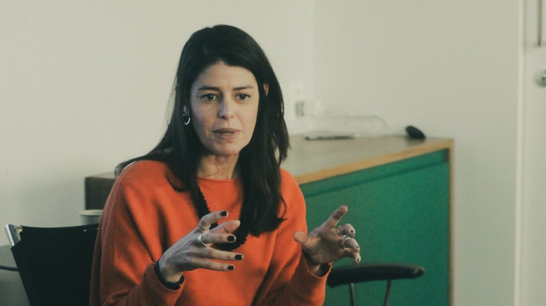 Imagem de Mariana Almeida. Ela é uma mulher branca, tem cabelo na tonalidade castanho-escuro e usa uma blusa laranja. Ela aparece gesticulando enquanto fala durante o evento.