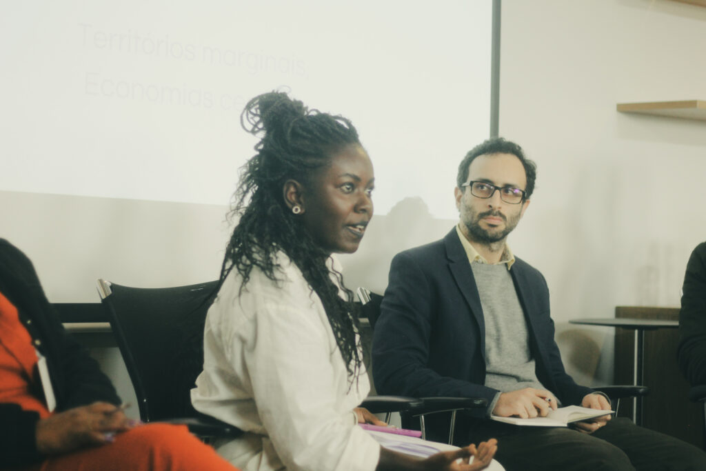 Imagem de Kenia Cardoso. Ela é uma mulher negra e usa tranças em formato box braid. Ela está com uma camisa na cor branca.