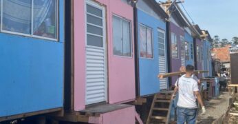 Imagem de mutirão do projeto +Lapena Habitar. A imagem mostra casas construídas durante a atividade, nas cores azul-claro, rosa e roxo, e pessoas voluntárias carregando uma viga de madeira.