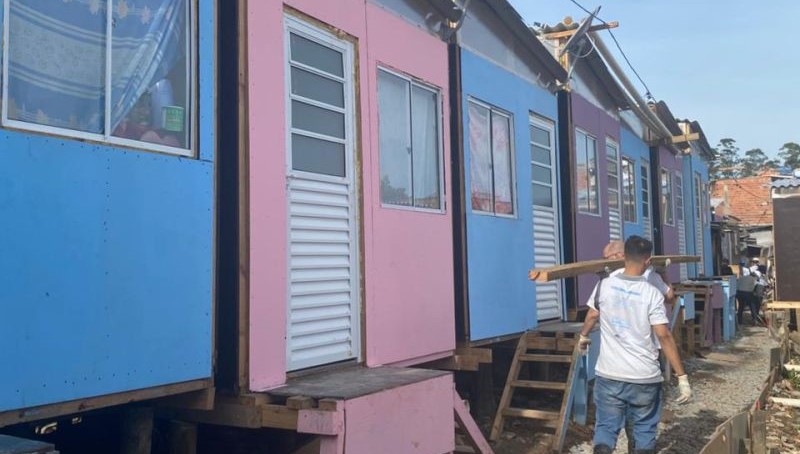 Imagem de mutirão do projeto +Lapena Habitar. A imagem mostra casas construídas durante a atividade, nas cores azul-claro, rosa e roxo, e pessoas voluntárias carregando uma viga de madeira.