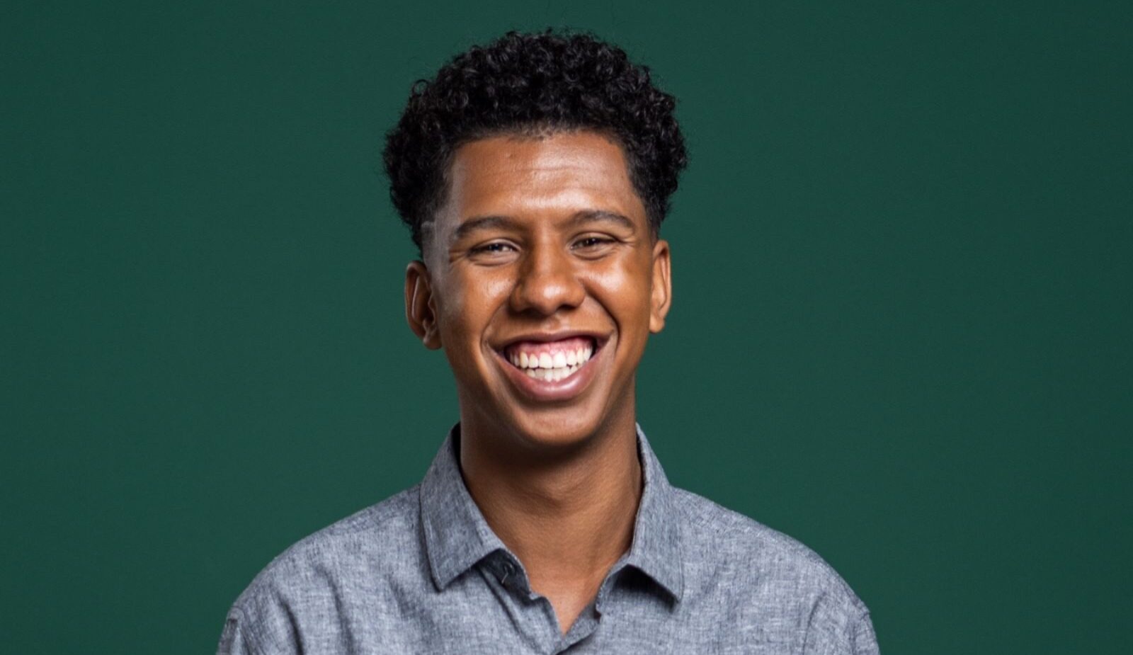 Imagem de Allan Anjos Dantas. Ele é um homem negro. Allan tem black power - com cachos - e aparece sorrindo. Ele usa uma camisa cinza e está à frente de um fundo verde.