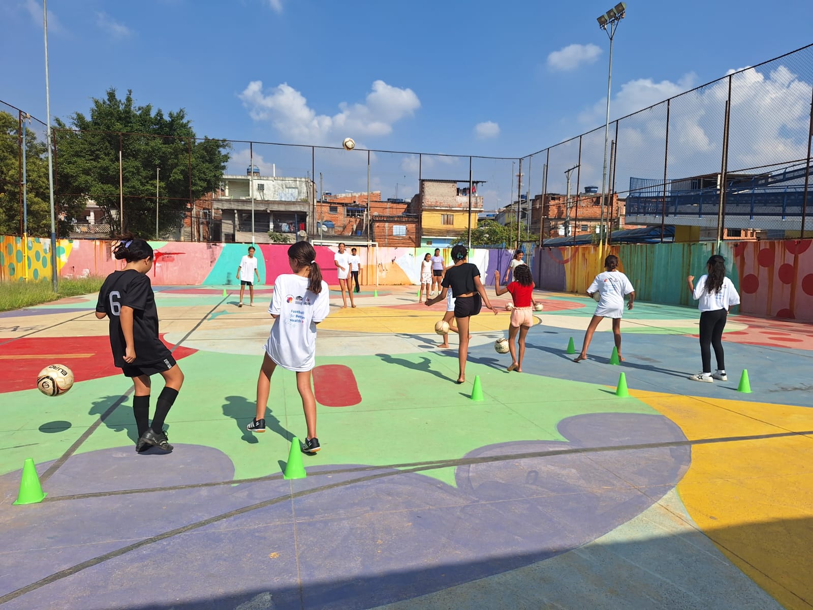 Imagem de treino do projeto Lapenna para Mulheres. As meninas aparecem jogando futebol em uma quadra multicolorida, na qual se destacam formas abstratas nas cores lilás, verde, laranja e azul.
