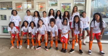 Imagem de integrantes do projeto Lapenna para Mulheres. Há 17 meninas usando o uniforme da iniciativa (calção e meiões laranja e camisa branca). Elas estão à frente de uma parede com fotografias.