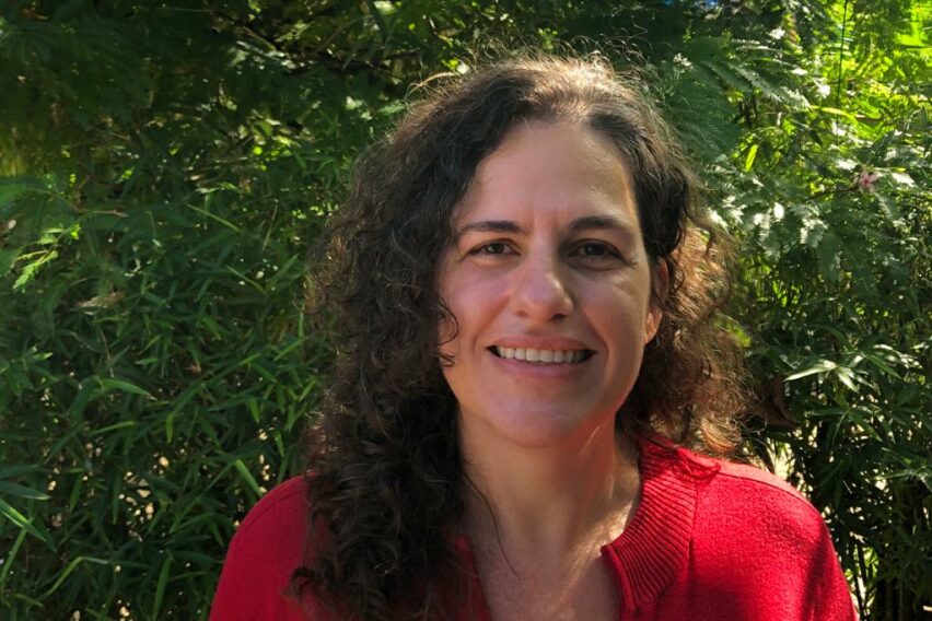 Imagem de Ludmila Costhek Abílio. Ela é uma mulher branca, com cabelos ligeiramente cacheados. Ludmila aparece sorrindo, usando uma blusa vermelha e à frente de fundo composto por plantas.