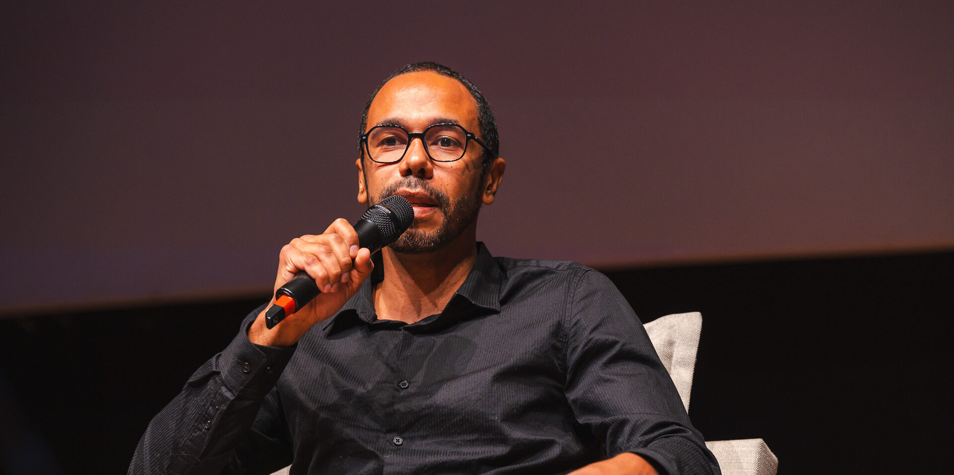 Imagem de Alan Pessoa Valadares. Ele é um homem negro, tem cabelo curto e usa óculos, camisa cinza e calça bege. Ele está sentado no palco de um auditório e fala ao telefone.