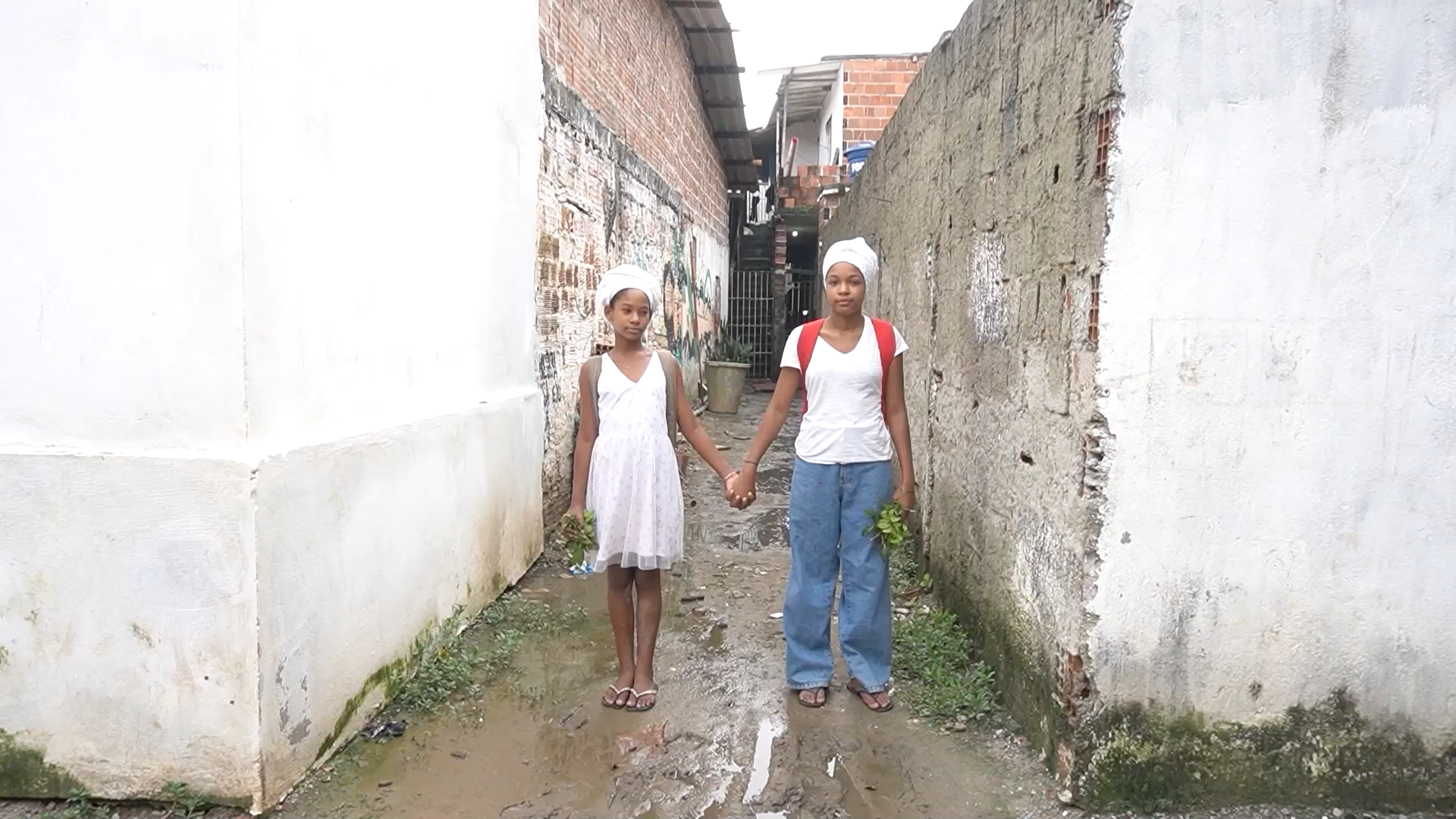 Imagem de duas garotas de mãos dadas em meio a um beco. Uma delas usa um vestido branco e a outra, uma camiseta branca e calça jeans - ambas usam lenços na cabeça na cor branca. A foto é uma cena do curta-metragem 'Orí Semente'.