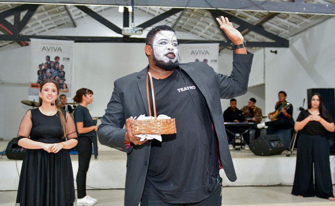 Intervenção teatral durante o Aviva Lapenna. Há um homem ao centro que está com maquiagem na cor branca e usa camiseta e blazer pretos - ele segura uma caixa com a mão direita e está com a mão esquerda erguida. Além dele, outras pessoas aparecem ao fundo.