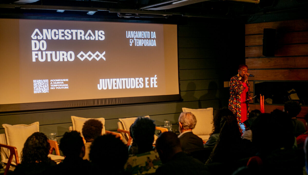 Imagem do auditório do Itaú Cultural. Aparece ao centro a tela com o logo do Ancestrais do Futuro e as demais informações sobre o evento. Pode-se ver também o público, assim como Mana Bella, a mestre de cerimônias, à direita.