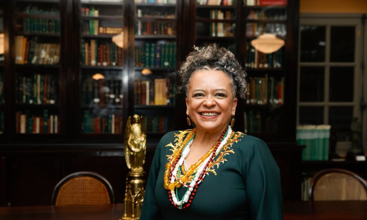 Imagem de Ana Maria Gonçalves. Ela aparece com um traje relativo à Academia Brasileira de Letras, sorrindo e com cabelos encaracolados e grisalhos presos no topo da cabeça. Ela aparece à frente de uma estante fechada com livros.