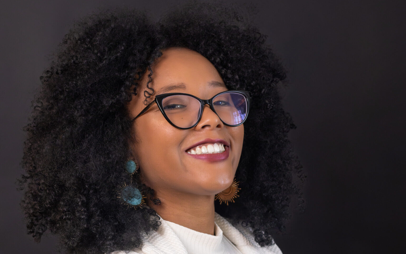 Imagem de Maíra Rodrigues da Silva. Ela tem cabelos cacheados e crespos. Maíra usa óculos, blazer e blusa brancas e aparece sorrindo à frente de um fundo escuro.