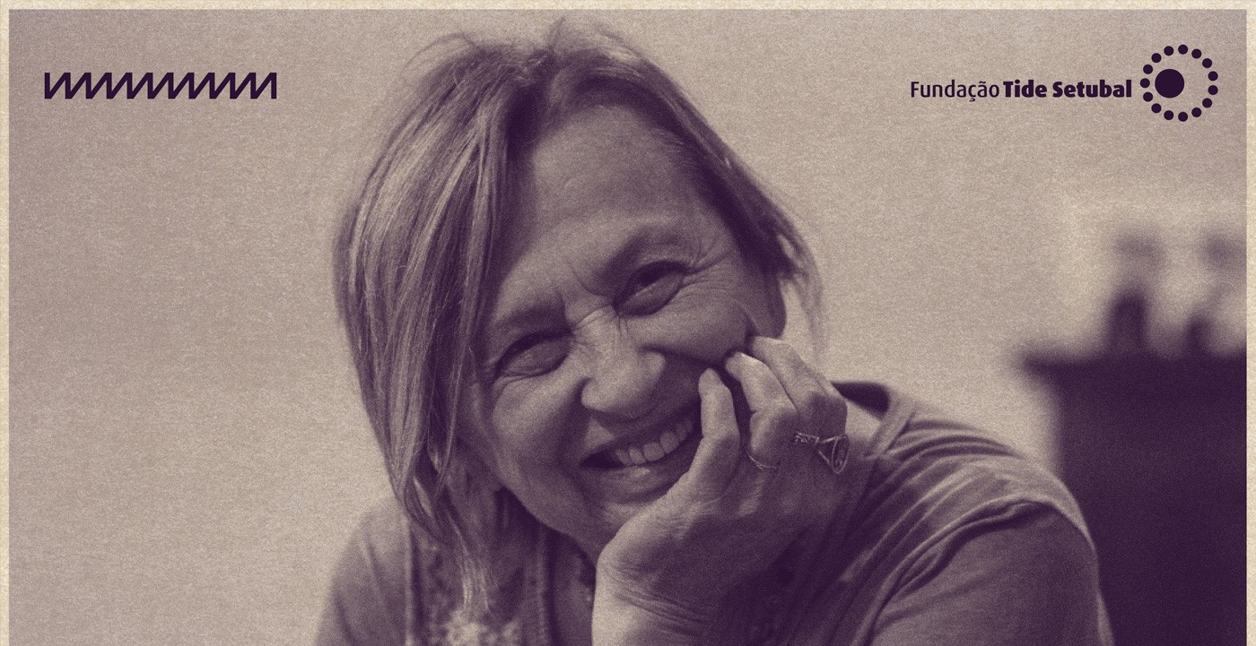 Imagem de Schuma Schumaher. Ela é uma mulher branca com cabelos loiros. Ela aparece sorrindo e com a mão esquerda apoiando o queixo. A foto tem filtro na cor roxa e os logos da Rádio Novelo e da Fundação Tide Setubal.