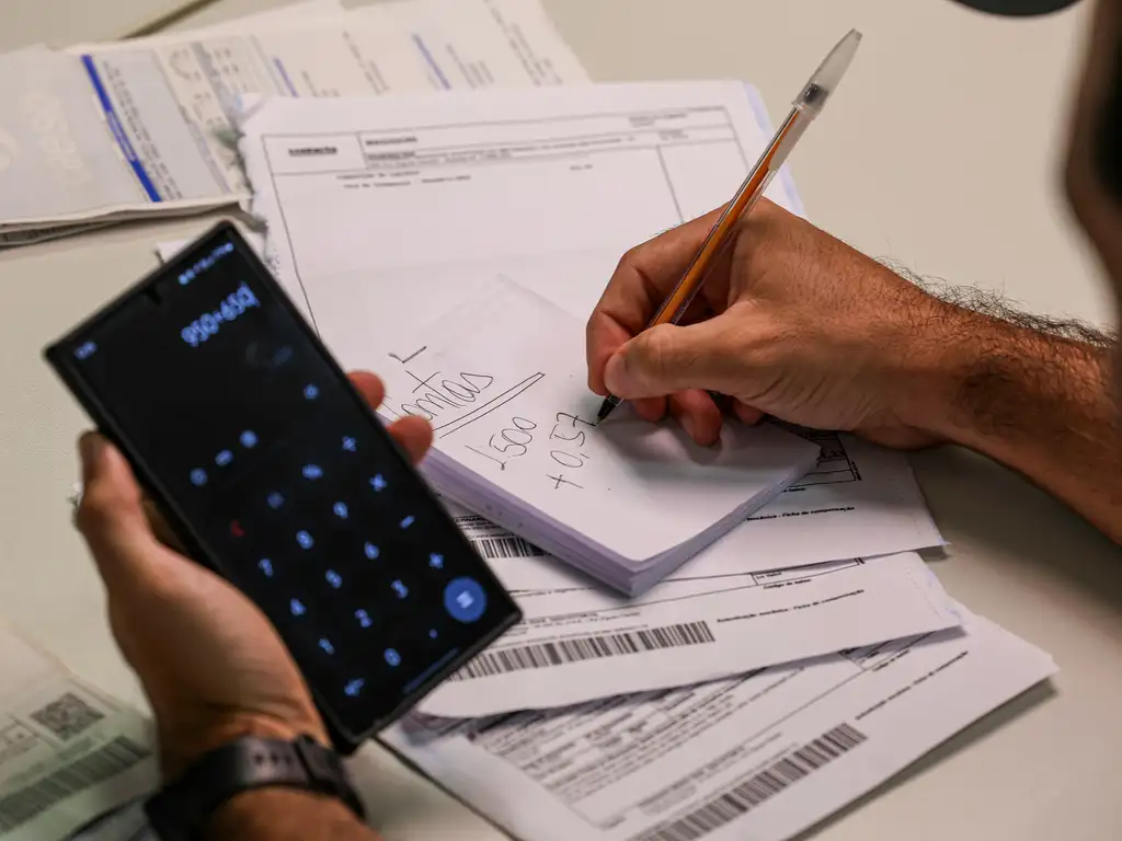 Imagem de uma pessoa segurando um smartphone, com o aplicativo de uma calculadora, na mão esquerda, enquanto faz anotações em um bloco com a mão direita. Há, sobre a mesa que essa pessoa usa, boletos relativos a contas diversas. A foto ilustra a nota sobre estrutura tributária.