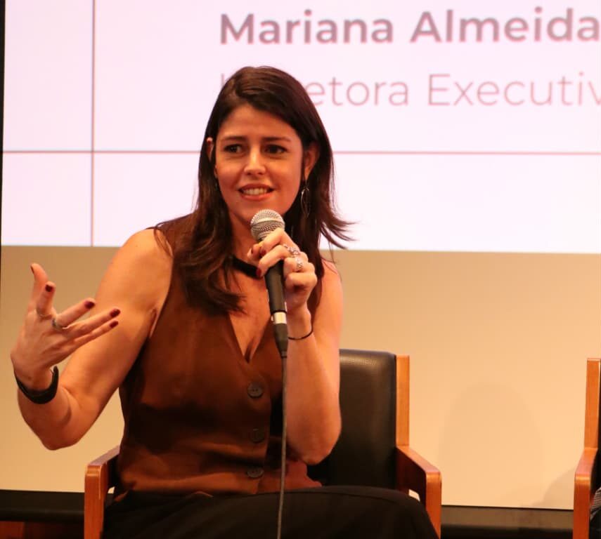 Imagem de Mariana Almeida. Ela tem cabelo castanho escuro e usa colete marrom e calça preta. Ela fala para pessoas em um anfiteatro e usa um microfone.