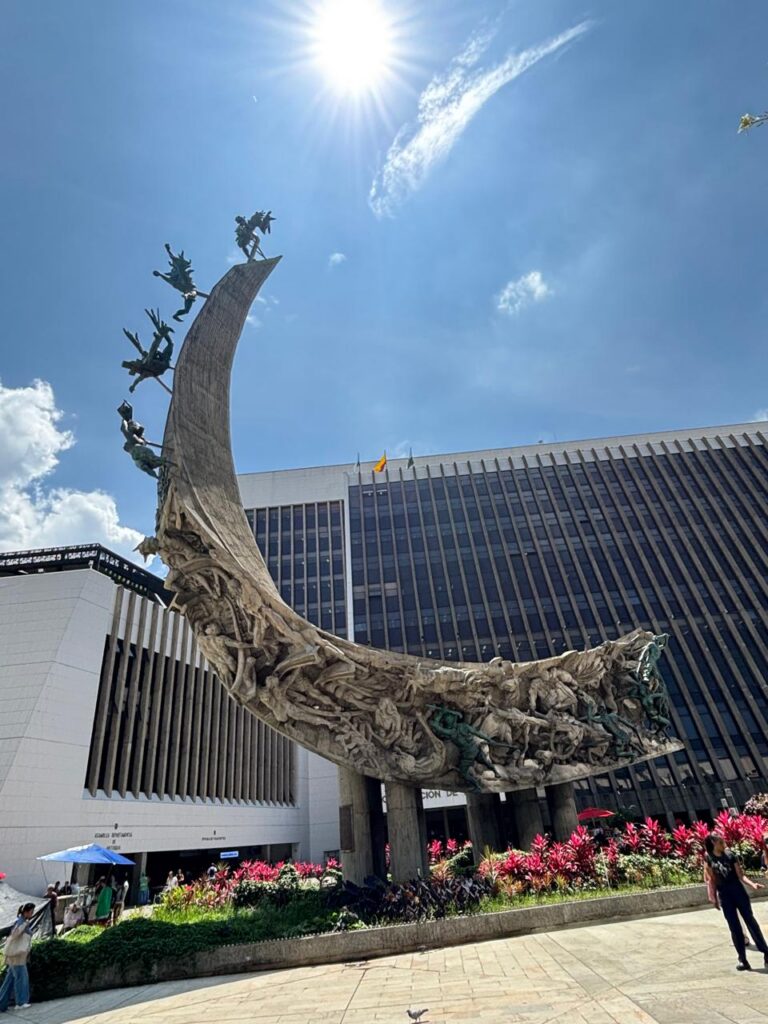 Imagem do Centro Administrativo La Alpujarra. A foto mostra dois prédios - um no canto esquerdo e outro ao fundo. Há um monumento em formato curvado com elementos inseridos no topo até a base.