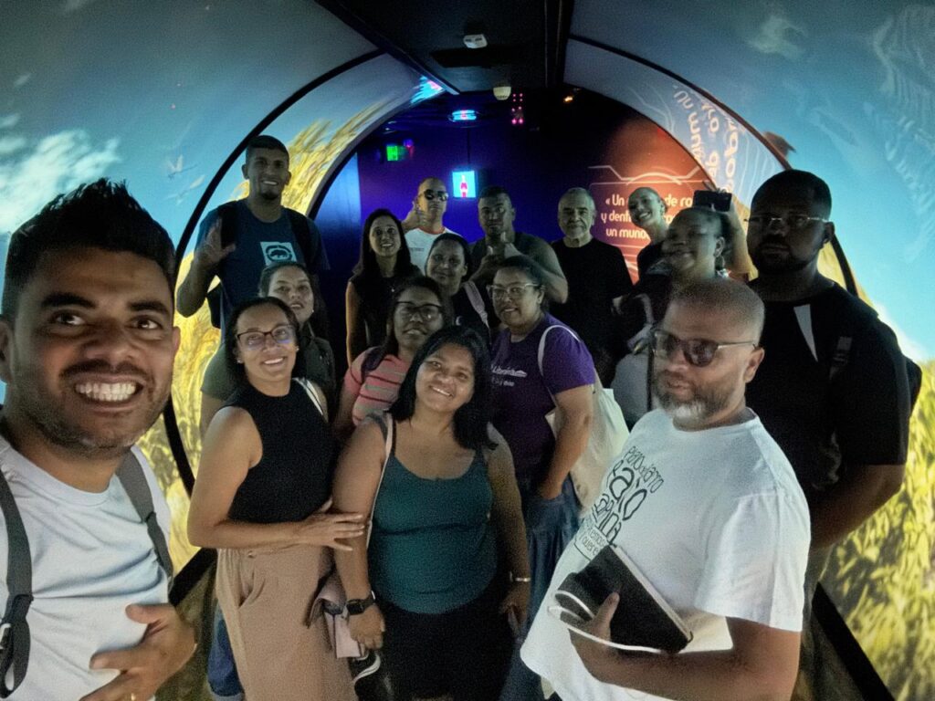 Um grupo de pessoas está dentro do Museu da Água. Eles aparecem sob uma abóbada branca e à frente de uma parede colorida com predominância de tonalidades azuis e terrosas. Trata-se do grupo que fez parte da viagem de imersão a Medellín.