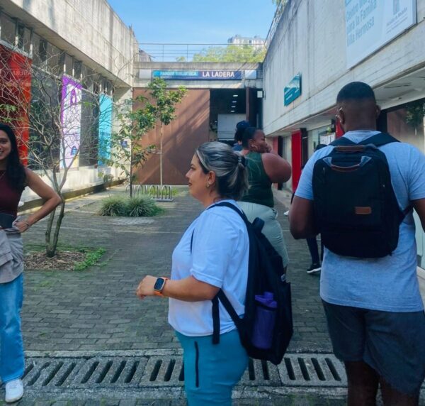Fachada do Parque Biblioteca La Ladera. Trata-se de uma construção com dois andares com três prédios conectados entre si. Há, ao centro, uma placa com o nome do espaço e pode-se ver, mais à frente, um pátio onde há três pessoas que participaram da viagem de imersão a Medellín.