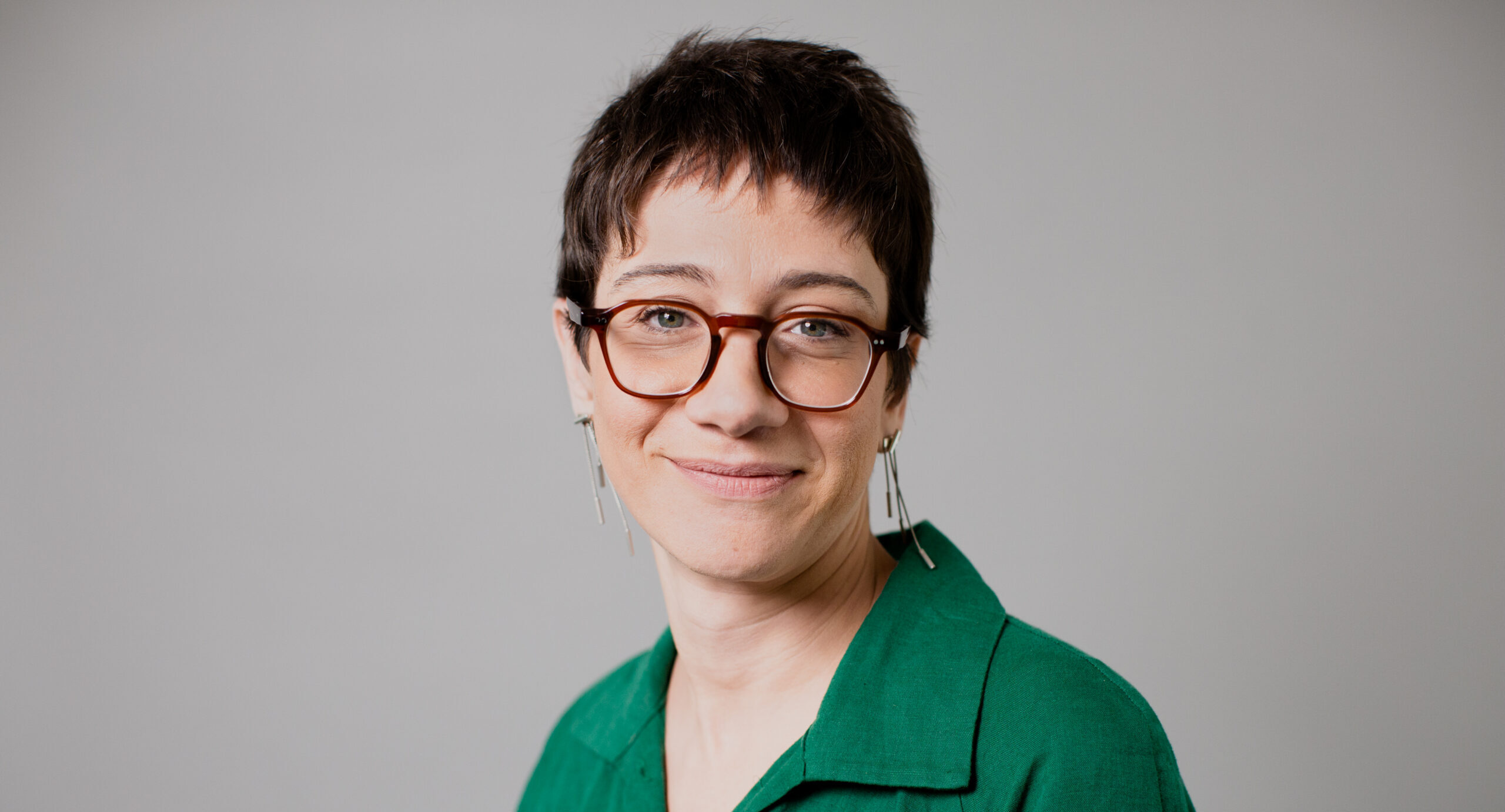 Imagem de Maria Mello. Ela tem cabelos curtos e usa óculos de acetato. Ela está também com par de brincos e uma camisa na cor verde.