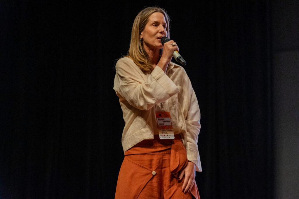 Imagem de Tide Setubal. Ela é uma mulher branca, com cabelos loiros e fala ao microfone. Ela usa uma blusa em tom ocre e calças com tonalidade alaranjada / terrosa.