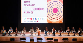 Representantes em um palco durante a abertura do Seminário Territórios Clínicos. Há, ao fundo, uma tela com a identidade visual do evento.