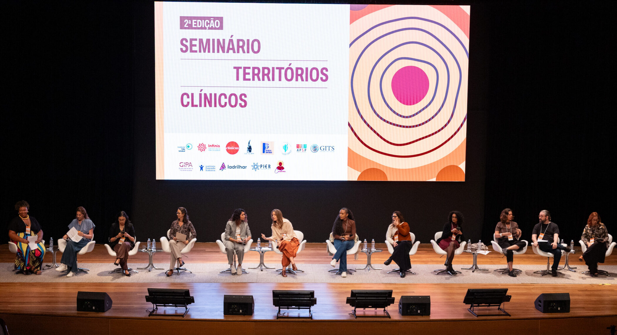 Representantes em um palco durante a abertura do Seminário Territórios Clínicos. Há, ao fundo, uma tela com a identidade visual do evento.