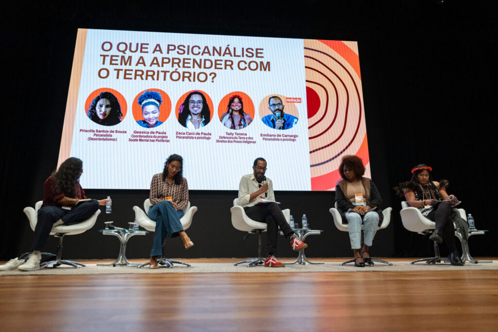 Imagem da mesa 'O que a psicanálise tem a ver com o território?'. As pessoas participantes estão no palco e há uma projeção de tela com o título da atividade em questão e as fotos das mesmas pessoas.
