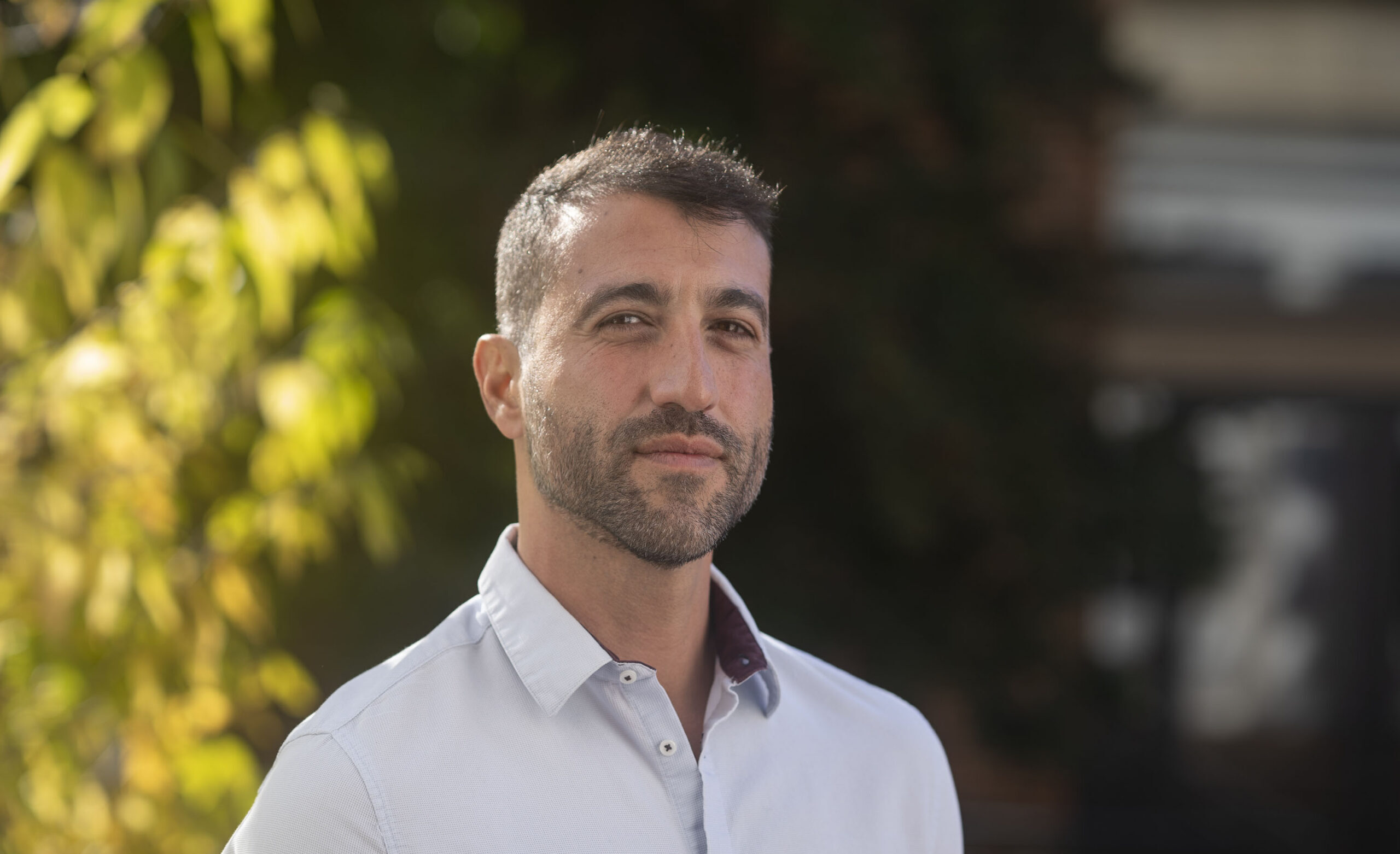 Imagem de David Nemer. Ele é um homem branco, tem barba e cabelo curto. Ele usa uma camisa na cor branca e está à frente de árvores.