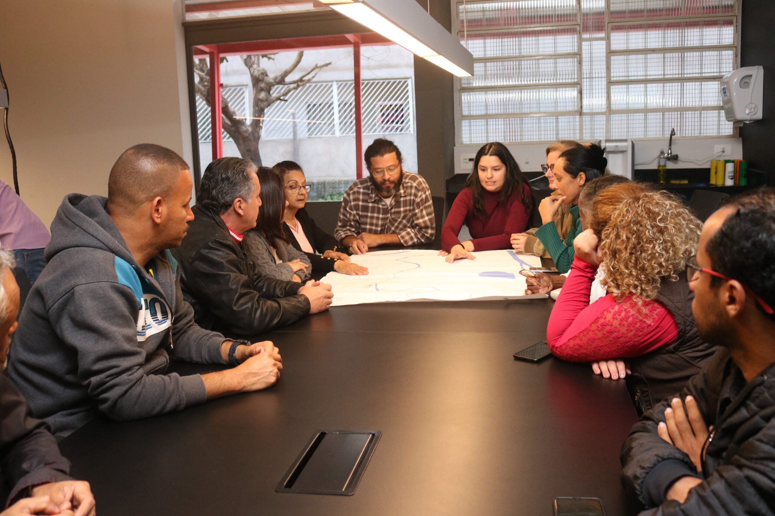 Imagem de integrantes do colegiado do Plano de Bairro do Jardim Lapena reunidos no coworking do Galpão ZL. Elas e eles estão ao redor de uma das mesas do espaço e analisam o mapa do bairro.
